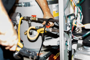 Plumber's hands working on yellow and black pipes connected to a water heater tank with red valve and internal wiring visible.