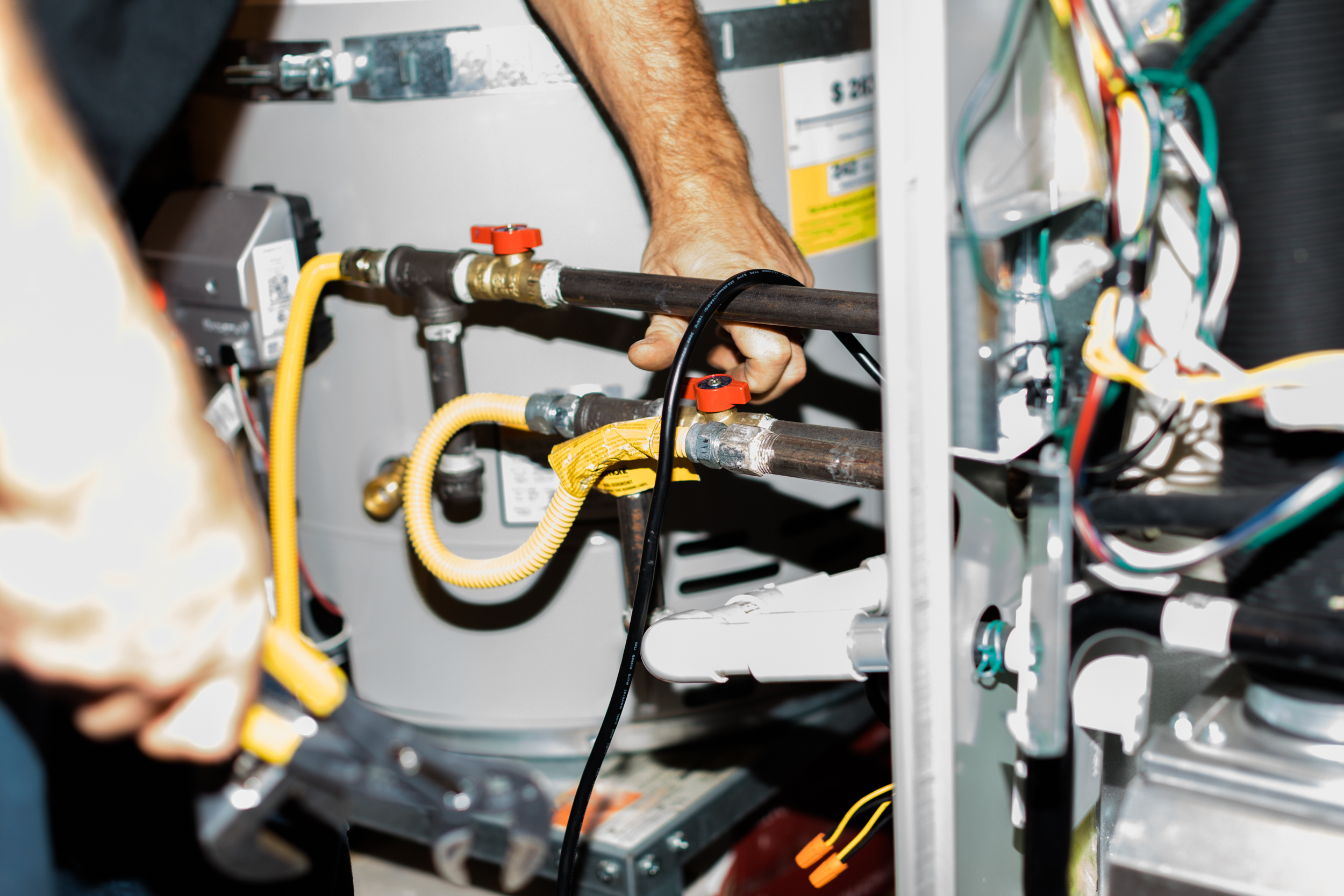 Plumber's hands working on yellow and black pipes connected to a water heater tank with red valve and internal wiring visible.