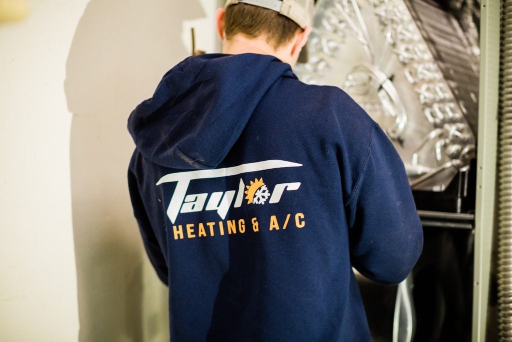 HVAC technician wearing a white cap and navy Taylor Heating & A/C hoodie working on heating system equipment indoors.