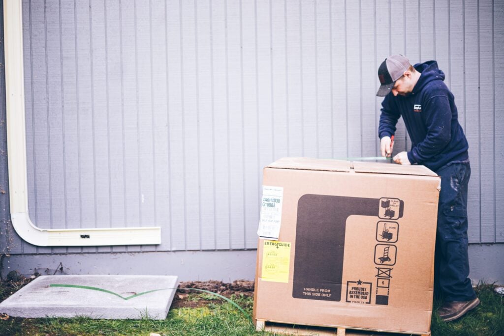 Person in navy hoodie and cap opens a large cardboard box with assembly instructions and an Energy Guide label against a white corrugated metal wall.