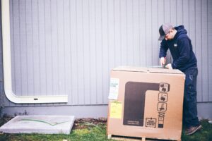 Person in navy hoodie and cap opens a large cardboard box with assembly instructions and an Energy Guide label against a white corrugated metal wall.
