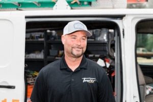 Professional tradesman in white cap and black Taylor Electric polo shirt standing in front of white service van with equipment visible in background.