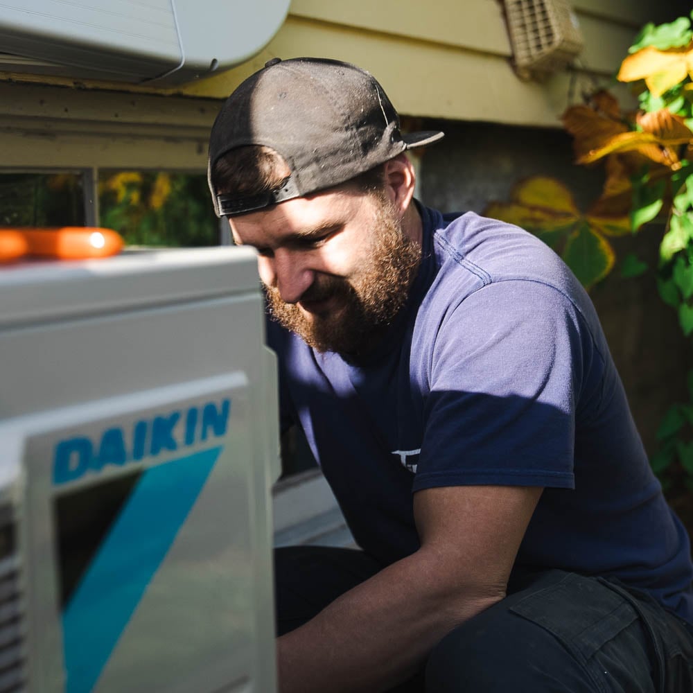 Bearded technician in blue shirt and cap inspecting a Daikin air conditioning unit outdoors near a residential home.