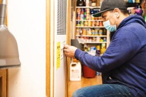 Service technician in blue hoodie and black cap wearing mask inspects refrigerator with yellow tag in appliance showroom.