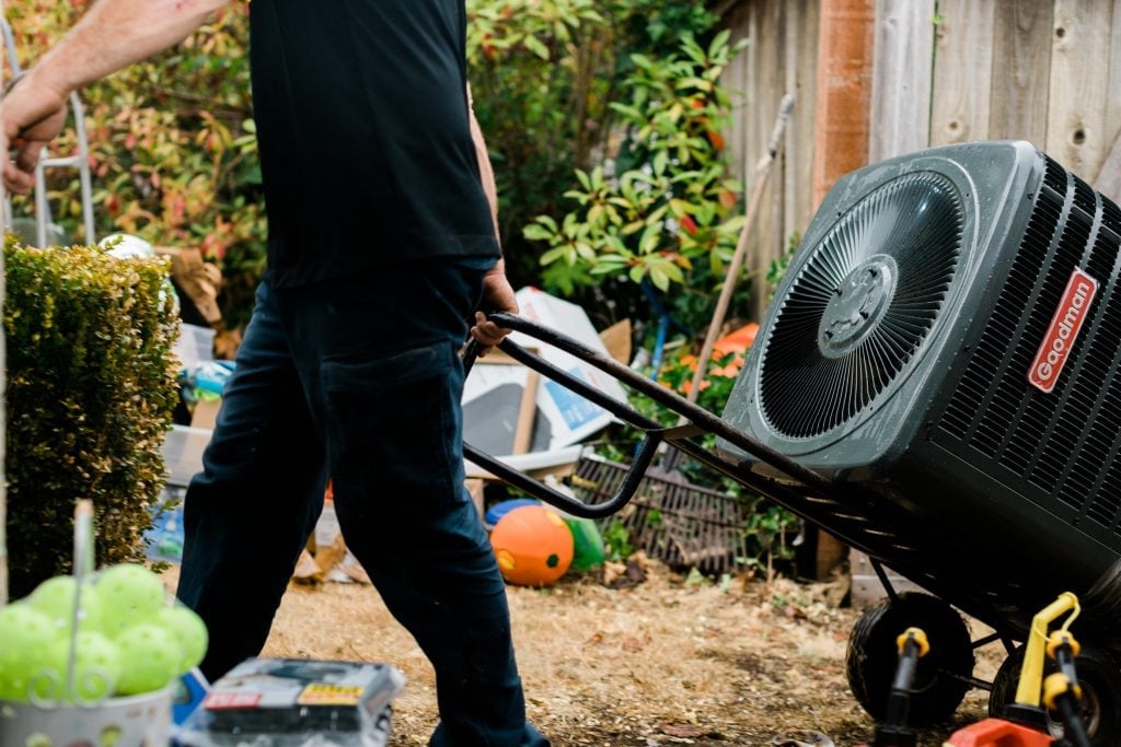 Person moving a Goodman air conditioning condenser unit on a hand truck in a residential backyard garden setting.