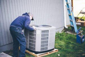 Technician in blue jacket and cap inspecting a white air conditioning condenser unit mounted on wooden platform against metal building siding.