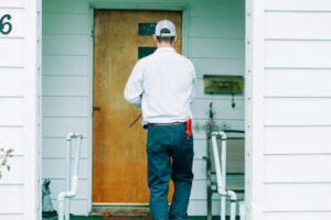 HVAC technician in white cap and shirt standing at wooden door numbered 6 of residential home with white siding.