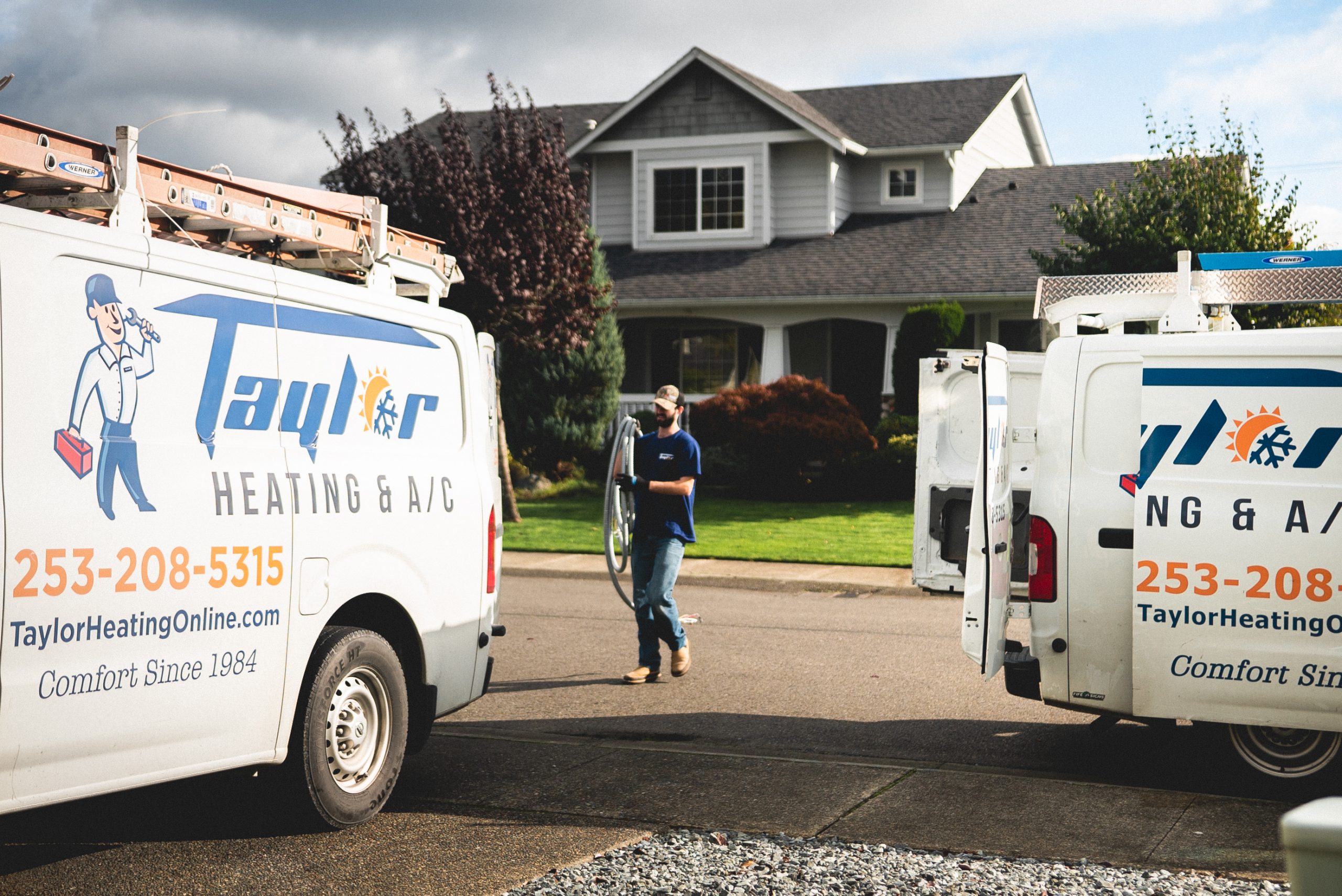 Two Taylor Heating & A/C service vans parked in a residential driveway with a technician standing between them near a two-story house.