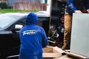 An HVAC technician in a blue branded jacket loads equipment into a service van in a residential driveway.