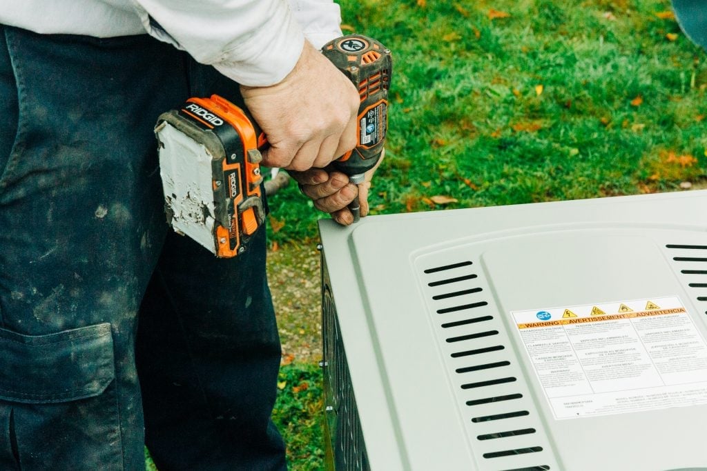 Technician using an orange and black cordless drill to work on a white HVAC unit outdoors with safety documentation visible.