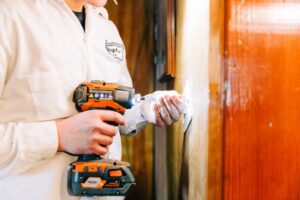 HVAC technician in white uniform holding orange and black thermal imaging camera against wall during inspection.