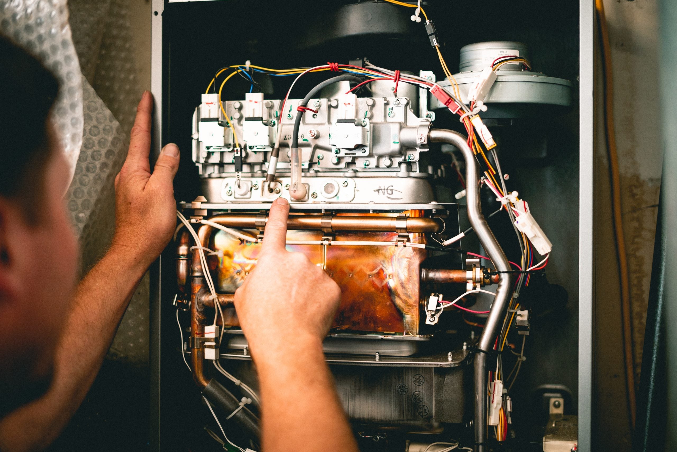 Technician pointing to internal components of an open furnace showing white switches, colored wiring, and copper heating elements.