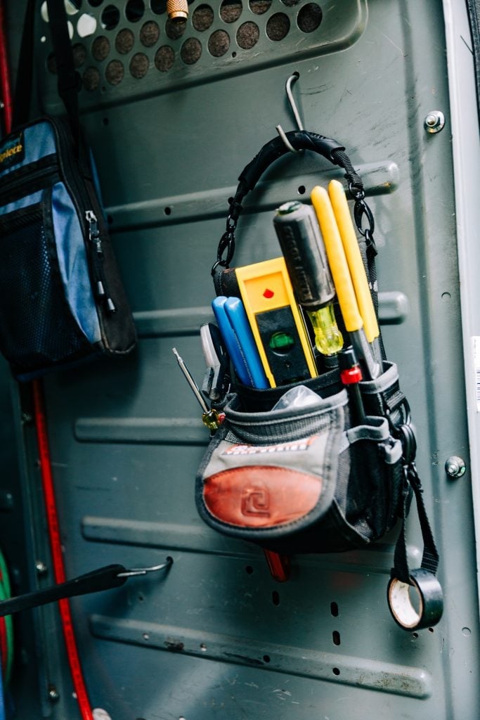 Gray tool bag with yellow and blue handles hanging on a green van interior wall with perforated metal storage panel.