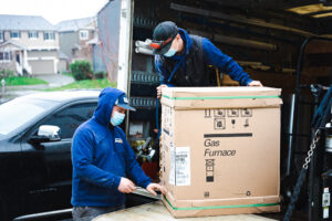 Two workers in blue jackets and masks unload a cardboard Gas Furnace box from the back of a delivery truck in a residential driveway.