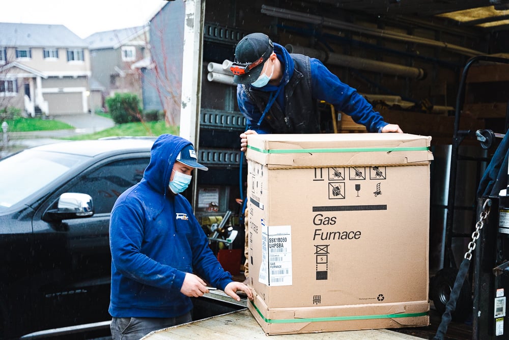 Two workers in blue jackets and masks unload a cardboard Gas Furnace box from the back of a delivery truck in a residential driveway.