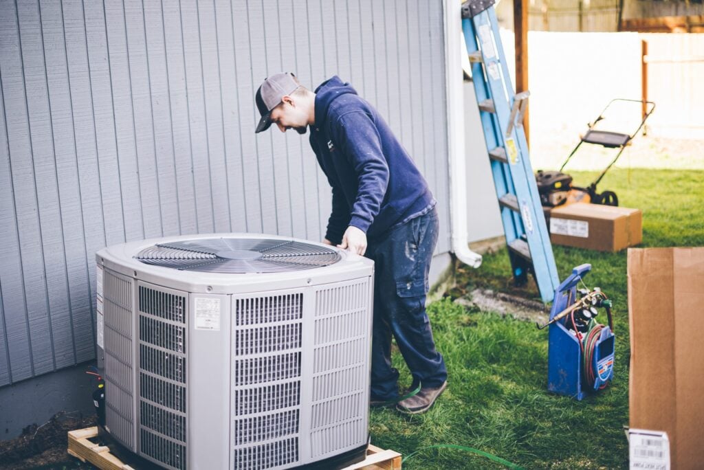 Technician in cap and blue jacket examining a white outdoor AC condenser unit mounted on grass near a metal shed.