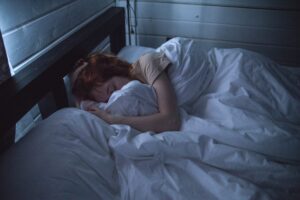 Person with reddish-brown hair sleeping on white bedding in a dimly lit bedroom at night.