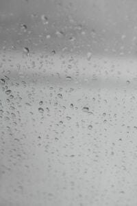 Multiple water droplets of varying sizes covering a wet glass surface against a gray sky background.