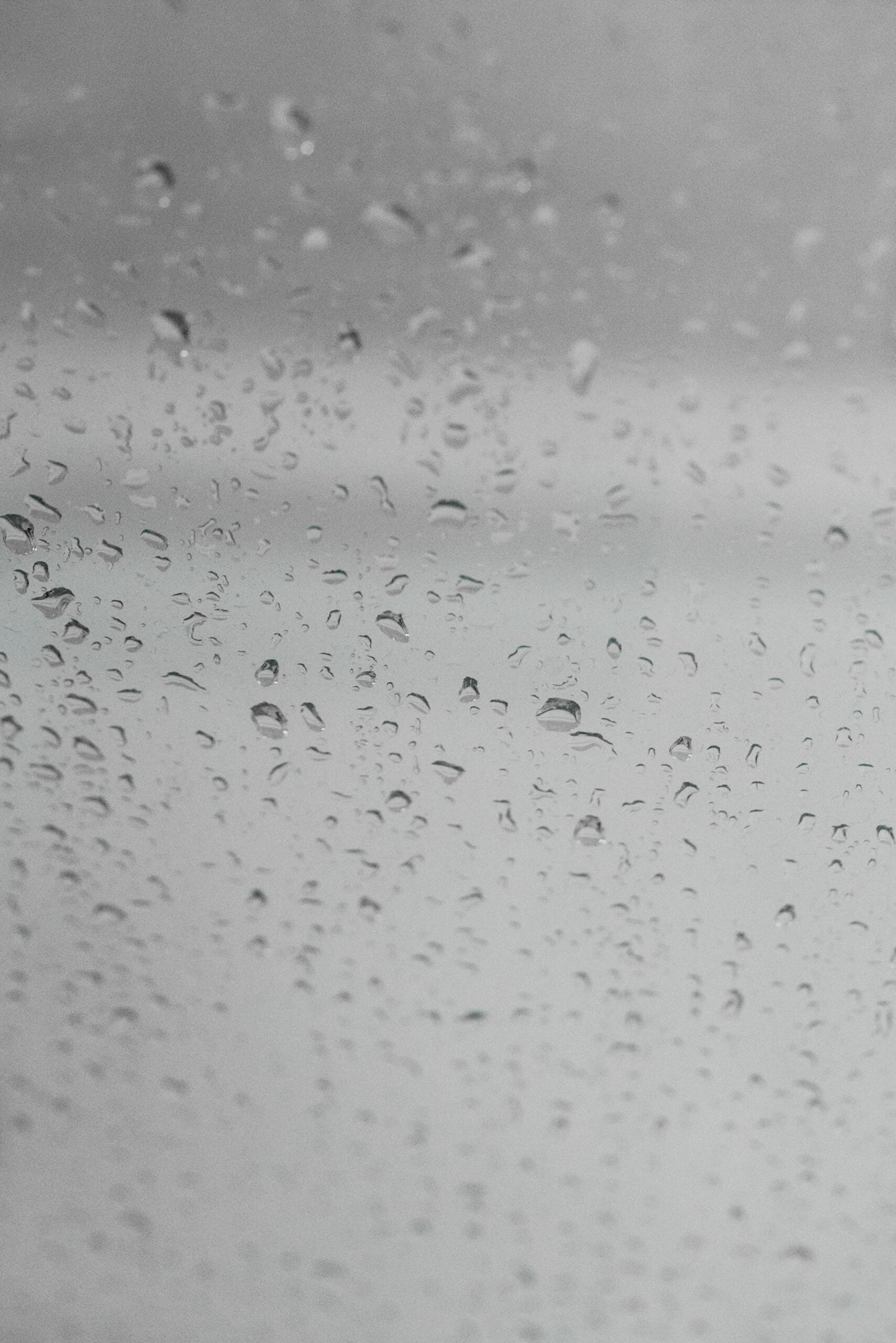 Multiple water droplets of varying sizes covering a wet glass surface against a gray sky background.