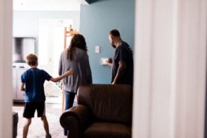 Young child in blue shirt stands with two adults in a home interior with blue-painted walls and brown furniture.