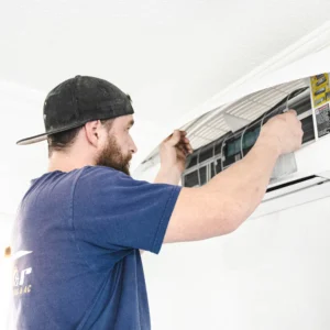 Technician in blue shirt and black cap replacing an air filter in a ceiling-mounted air conditioning unit.