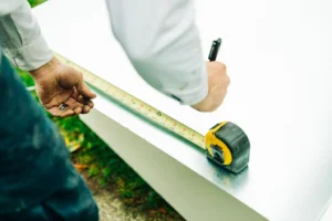 Technician using a yellow and black tape measure to measure white HVAC ductwork or equipment outdoors.