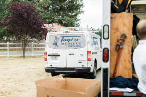 White Taylor Heating & A/C service van parked in residential driveway with equipment and tools visible in foreground.