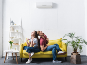 Young couple sitting together on a yellow sofa in a bright white room with an air conditioning unit mounted on the wall and potted plants nearby.
