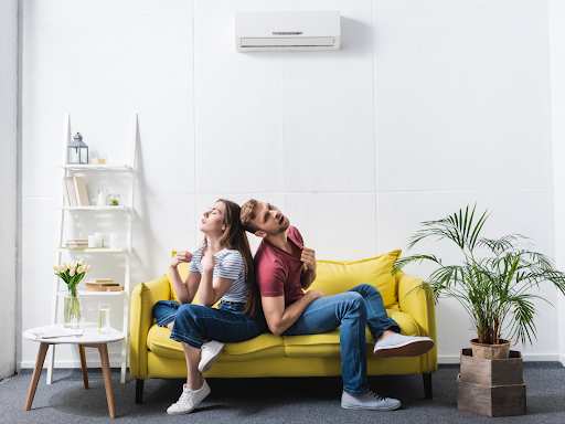 Young couple sitting together on a yellow sofa in a bright white room with an air conditioning unit mounted on the wall and potted plants nearby.