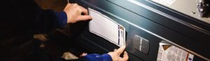 Person's hands examining a white pleated furnace air filter installed in a dark heating unit.