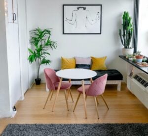 Bright modern living room features a white round table with pink chairs, black sofa with yellow pillows, potted plants, and line art on the wall.