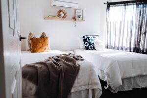 Bright, minimalist bedroom featuring two twin beds with white waffle-weave bedding, decorative pillows, and a brown throw blanket. Wooden floating shelf with su