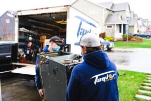 Two HVAC technicians in blue uniforms and caps load heating equipment into a mobile service truck in a residential neighborhood.