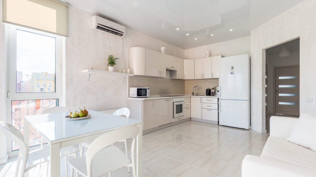 Bright modern kitchen featuring white cabinetry, stainless steel appliances, dining table with fruit bowl, and large window overlooking cityscape.