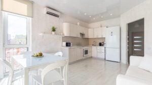 Bright modern kitchen featuring white cabinetry, stainless steel appliances, dining table with fruit bowl, and large window overlooking cityscape.