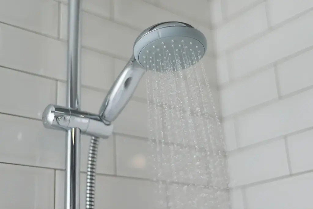 A chrome showerhead mounted on a vertical rail sprays water against white subway tile bathroom walls.