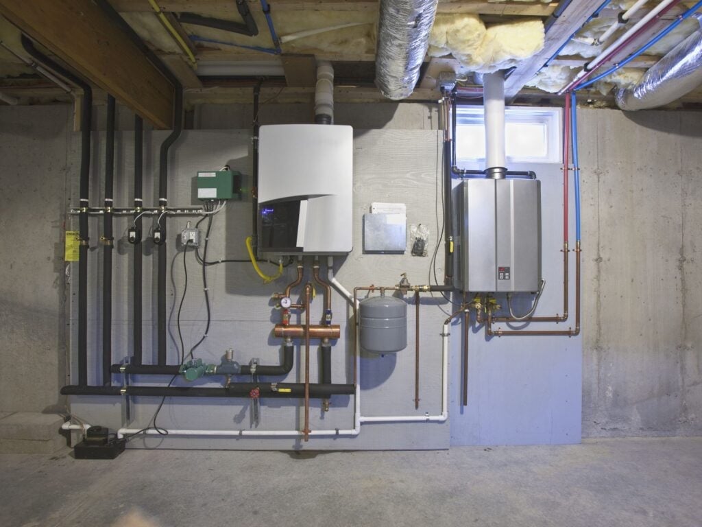 Modern tankless water heater system installed in basement with copper piping, expansion tank, and various mechanical components on concrete wall.