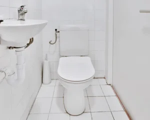 Minimalist white bathroom featuring a toilet with closed seat, pedestal sink with chrome faucet, and white tile flooring and walls.