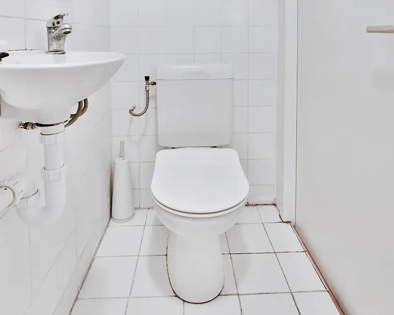 Minimalist white bathroom featuring a toilet with closed seat, pedestal sink with chrome faucet, and white tile flooring and walls.