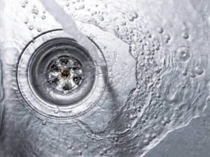 Close-up view of water swirling down a metal sink drain surrounded by bubbles and water droplets on a wet surface.