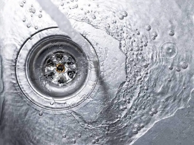 Close-up view of water swirling down a metal sink drain surrounded by bubbles and water droplets on a wet surface.