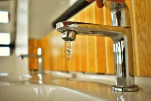 A modern chrome bathroom faucet with water dripping from the spout, mounted on a light marble sink countertop with wooden cabinetry visible in the background.