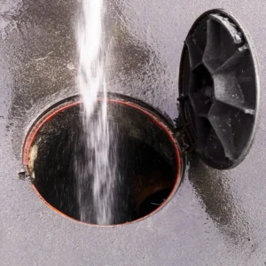 Water stream from hydrojetting equipment sprays forcefully into an open sewer manhole with red rim and black cover, cleaning pipes and removing debris.