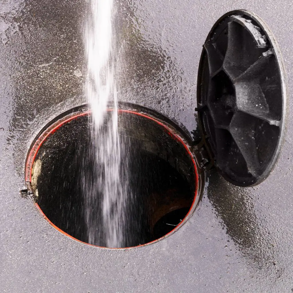 Water stream from hydrojetting equipment sprays forcefully into an open sewer manhole with red rim and black cover, cleaning pipes and removing debris.