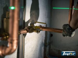 Green laser level beam aligned horizontally across copper pipes and plumbing fixtures on a concrete wall.