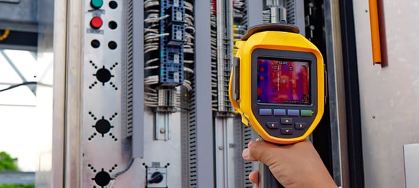 Technician holding a yellow and black thermal imaging camera displaying a heat map while inspecting an industrial electrical panel with wiring and circuit break
