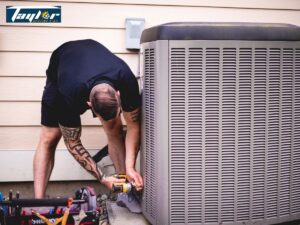 Technician in dark shirt bending over to service an outdoor air conditioning unit with tools nearby.