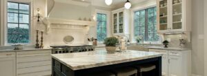Bright white kitchen featuring marble countertops, black gas range, white cabinetry with glass doors, and a black island with marble top.