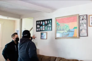 Two people in dark jackets stand in a living room discussing framed artwork on white walls, including family photos and landscape paintings.