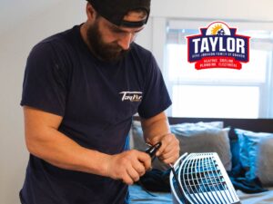 Technician wearing Taylor branded navy shirt and safety mask working on electrical components with tools indoors.